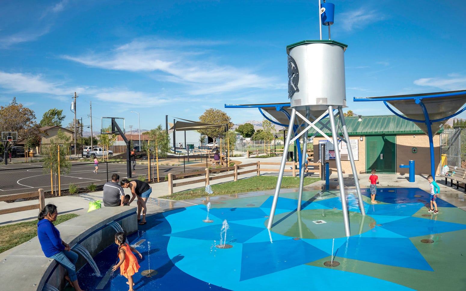 Splash pad with water tower that dumps water from a bucket and children playing—basketball courts in background